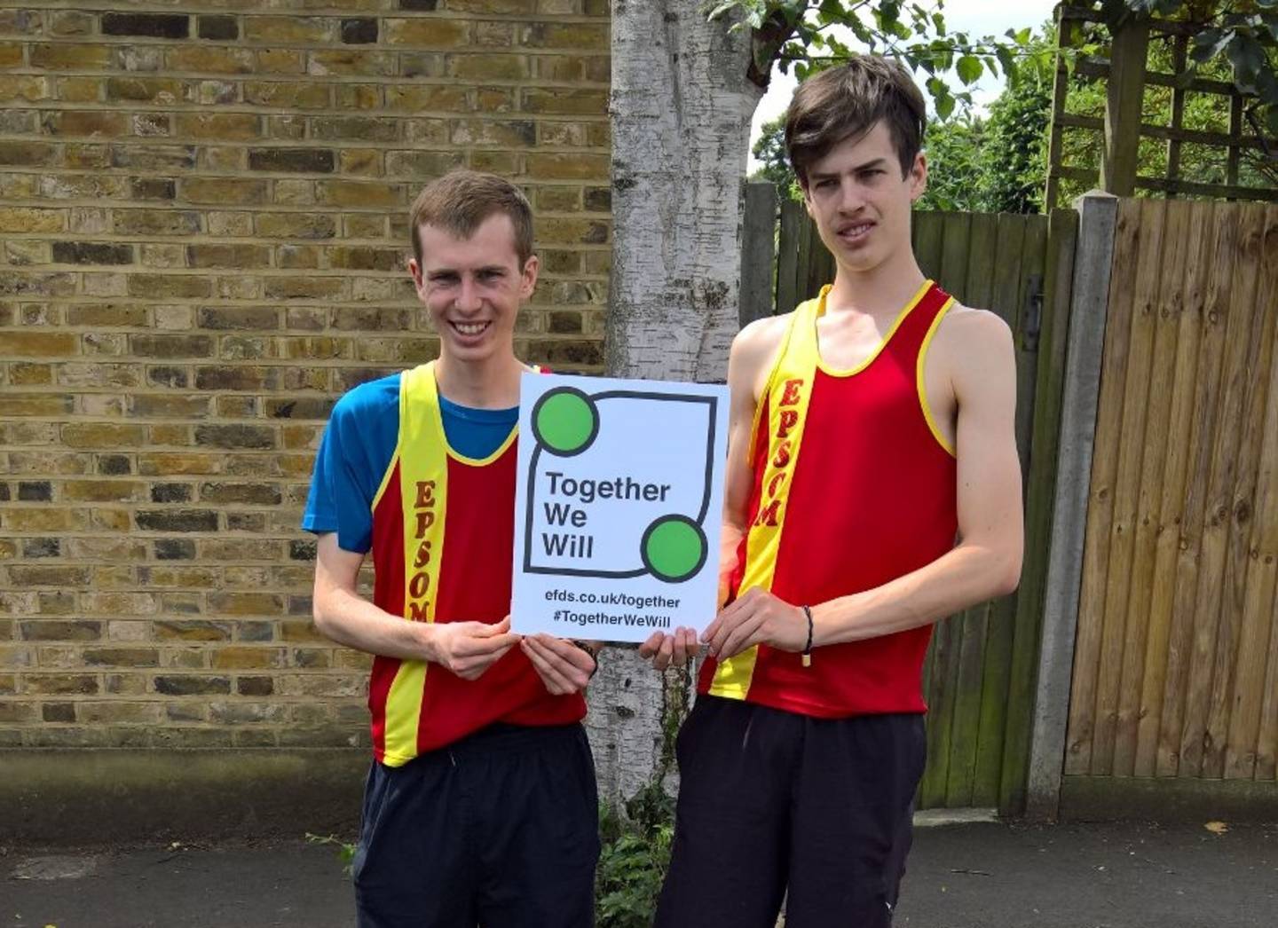 Image shows Daniel Wolff and team mate in their running kit smiling and holding a Together We Will campaign board.