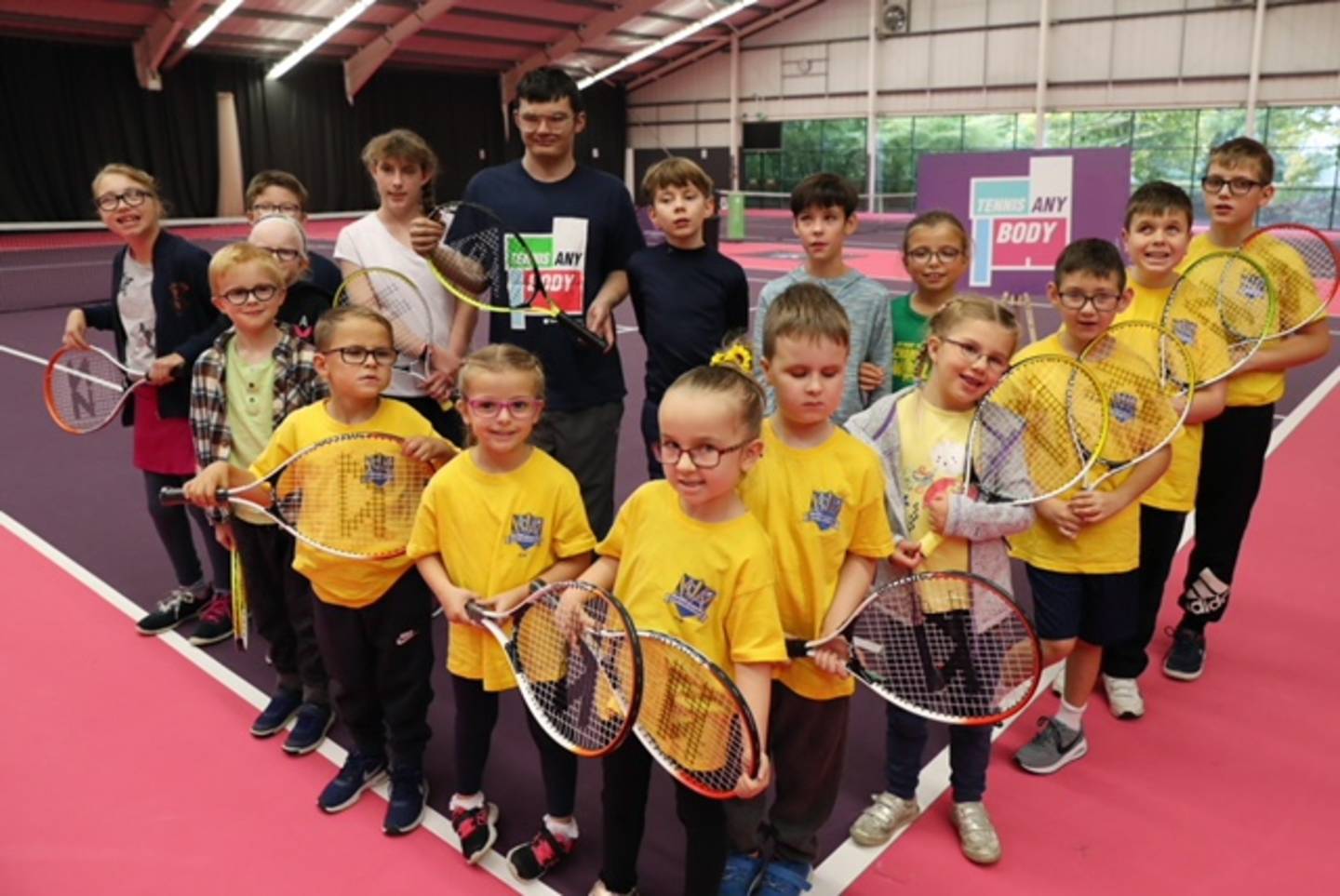 Group of children and tennis coach with tennis rackets in hand smiling to camera.