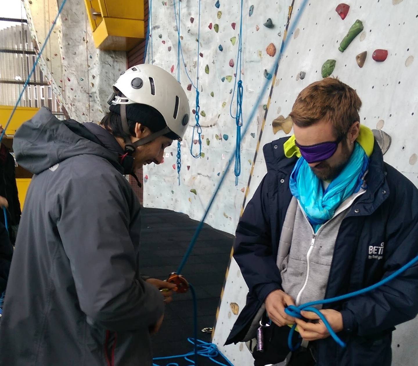 Two men, one blind folded, getting ready for an indoor climbing session. 