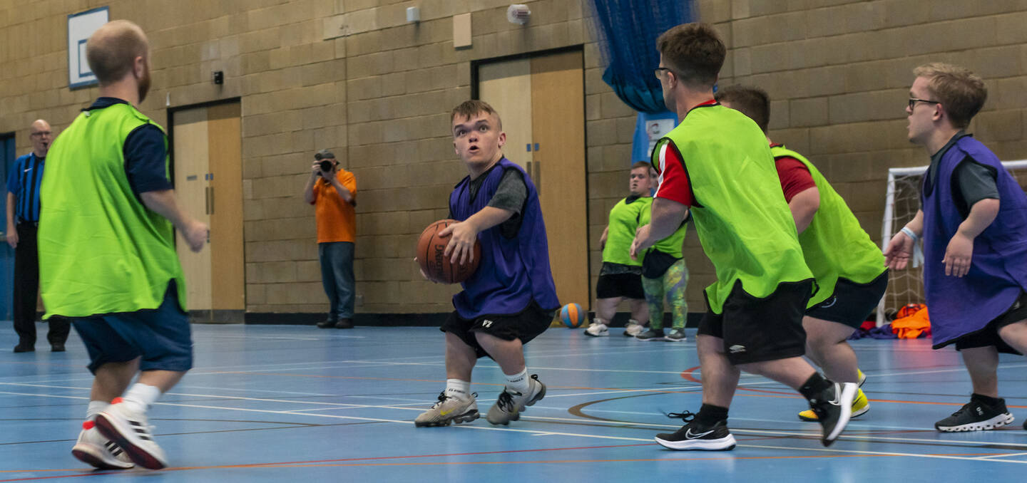 A man in a purple top holds a basketball as two people move towards him. 