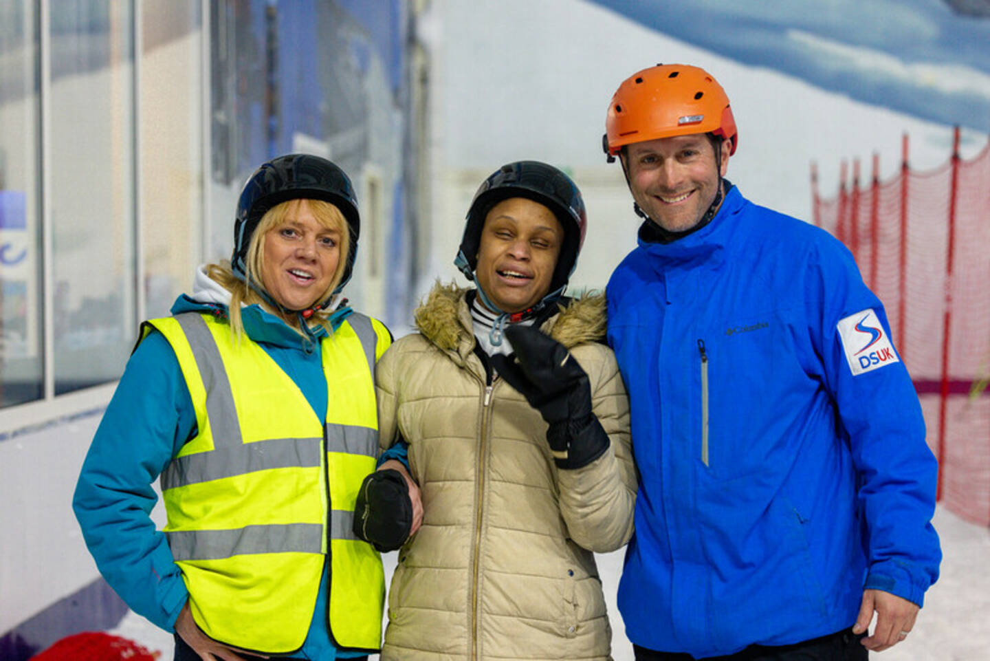 Two women, Tracey and Hazel, and a man, James, stand arm-in-arm at an indoor ski centre. They’re wearing thick coats and helmets and are smiling to camera.