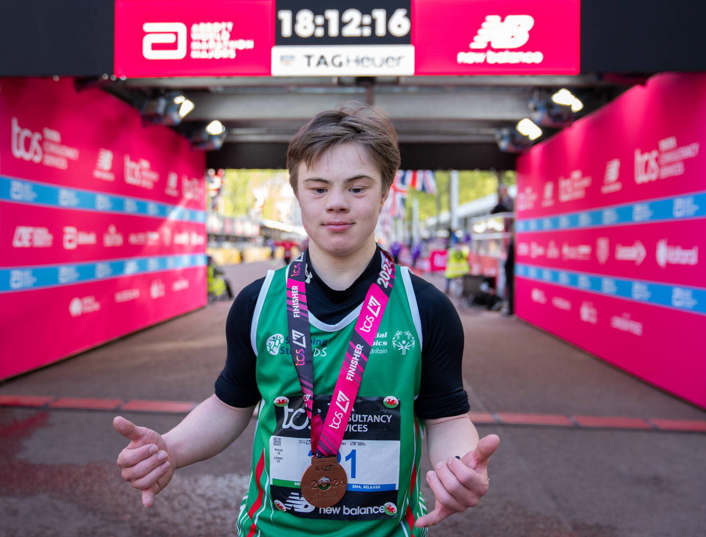Lloyd Martin, who has downs syndrome, is stood on the finish line of the London Marathon having just completed the race. He is wearing his medal and giving a thumbs up to the camera.