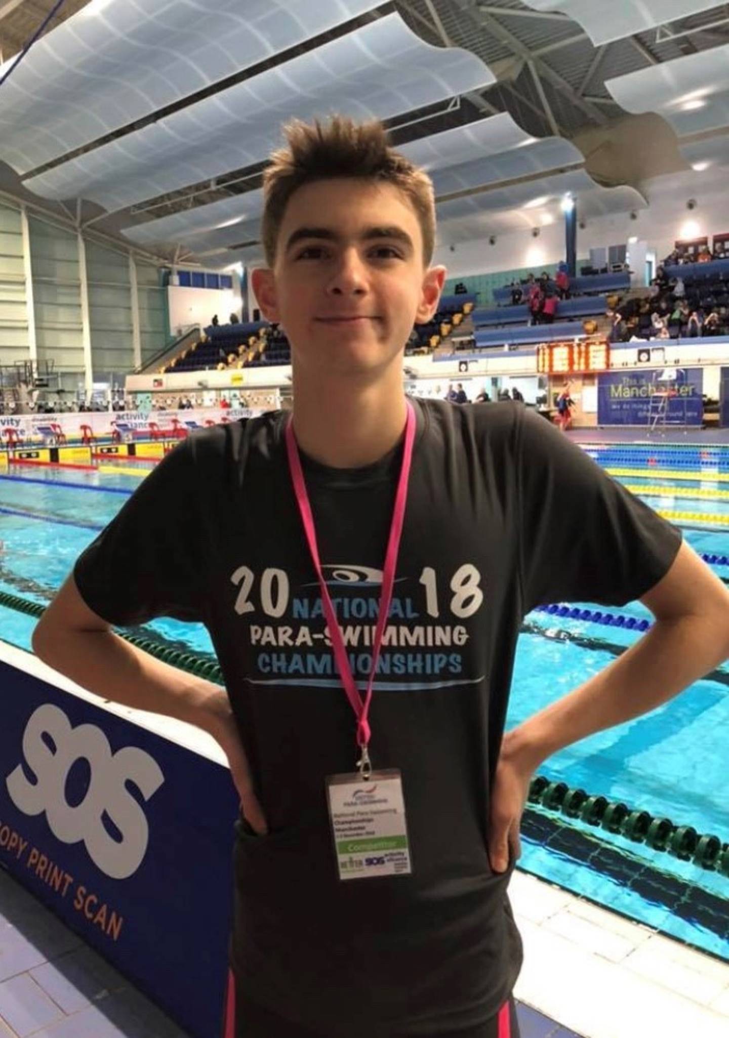 Joe smiling to camera at the Manchester National Swimming Championships in December 2018. Swimming pool is behind him. 