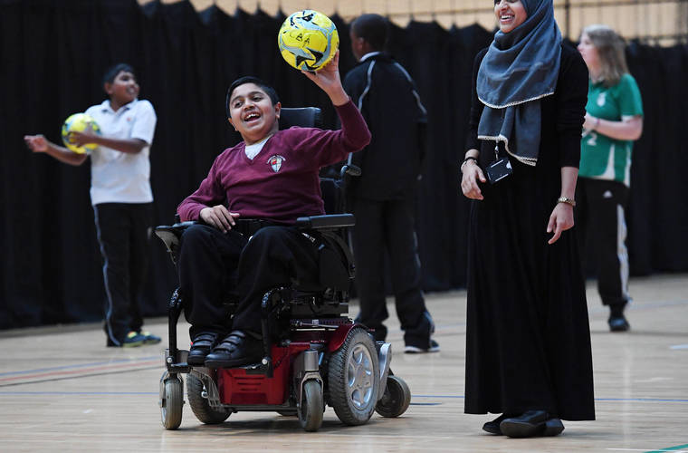 Boy in powerchair with football in hand laughing with girl stood next to him smiling. 