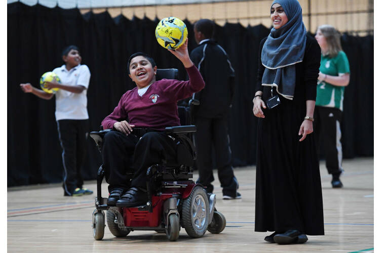 Boy in powerchair with football in hand laughing with girl stood next to him smiling. 