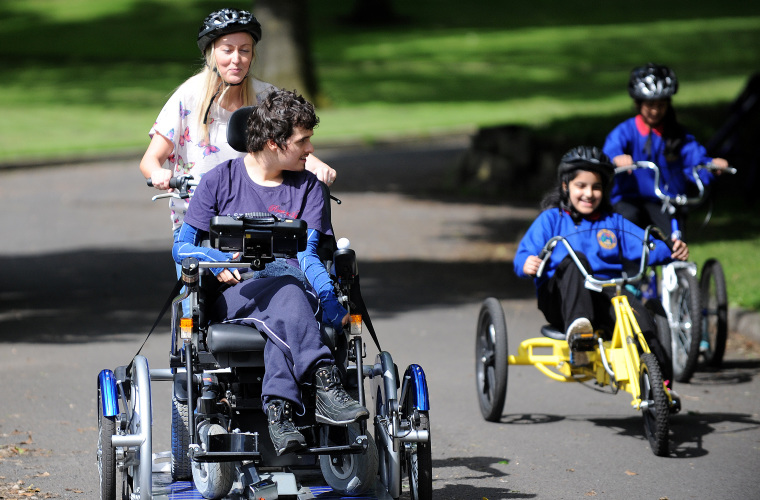 Young man riding adapted bike with supporter during a cycling session in the park  