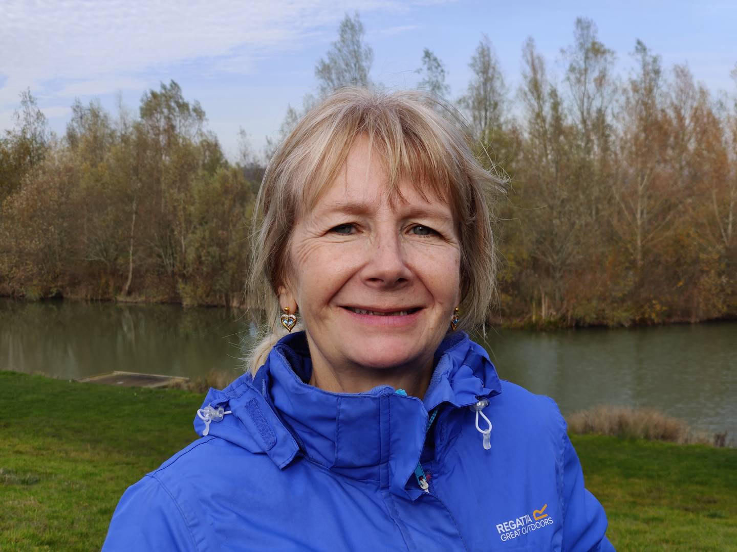 Shona outside with trees and a lake behind her smiling to camera. 