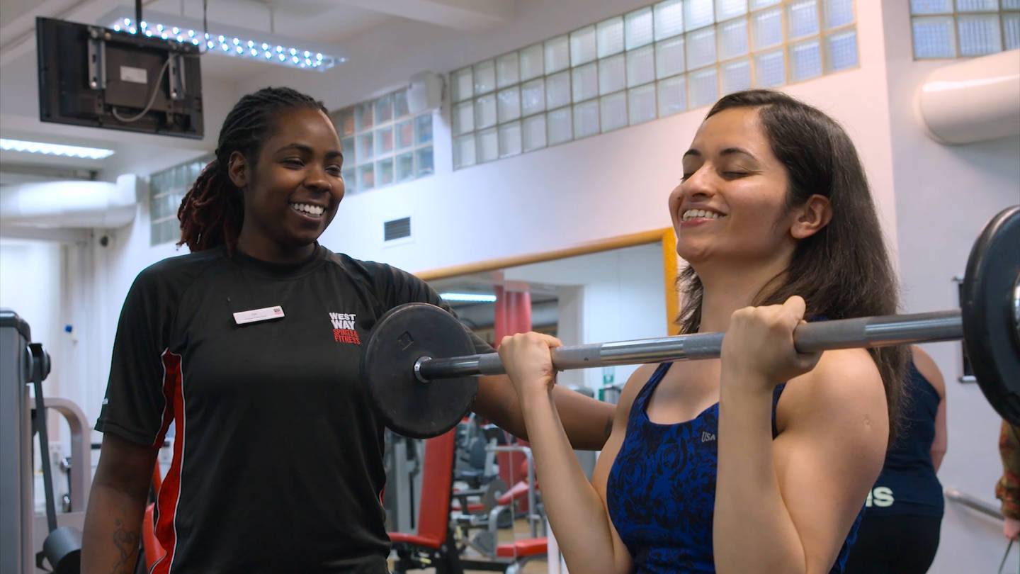 Visually impaired woman lifting barbell in the gym supported by a fitness instructor