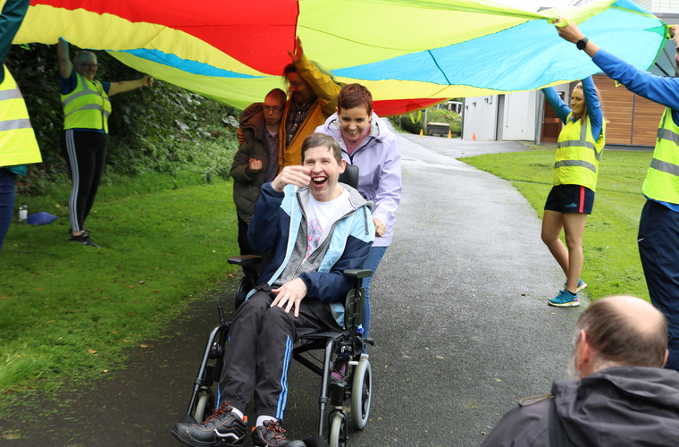 Wheelchair user smiling and laughing while going under a colourful parachute held up by volunteers 