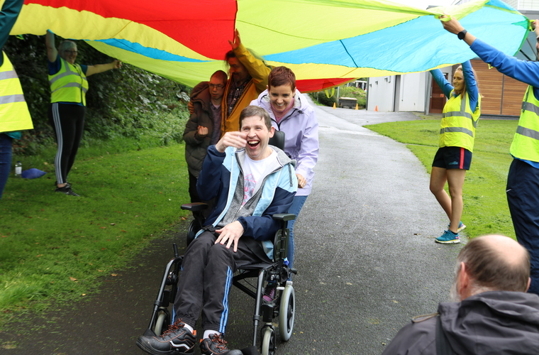 Wheelchair user smiling and laughing while going under a colourful parachute held up by volunteers