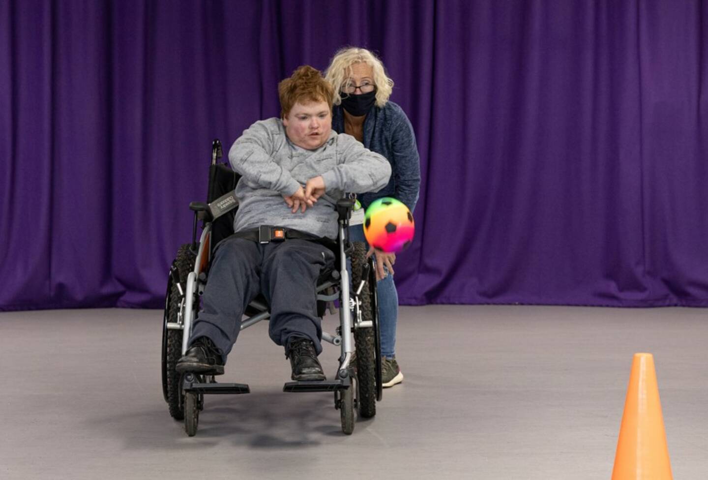 A man using a wheelchair propelling a multi-coloured ball with his hands, as a woman encourages close by.