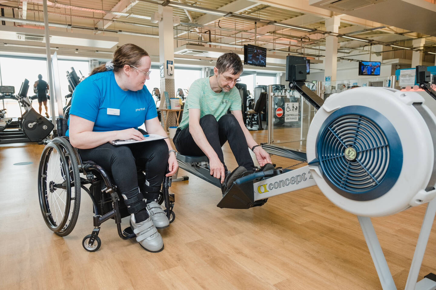 Man using the rowing machine in a gym with a woman personal trainer, who uses a wheelchair. 