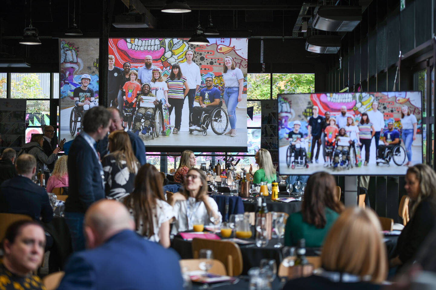 A room of people seated at tables with two screens showing an image of a group of people. 