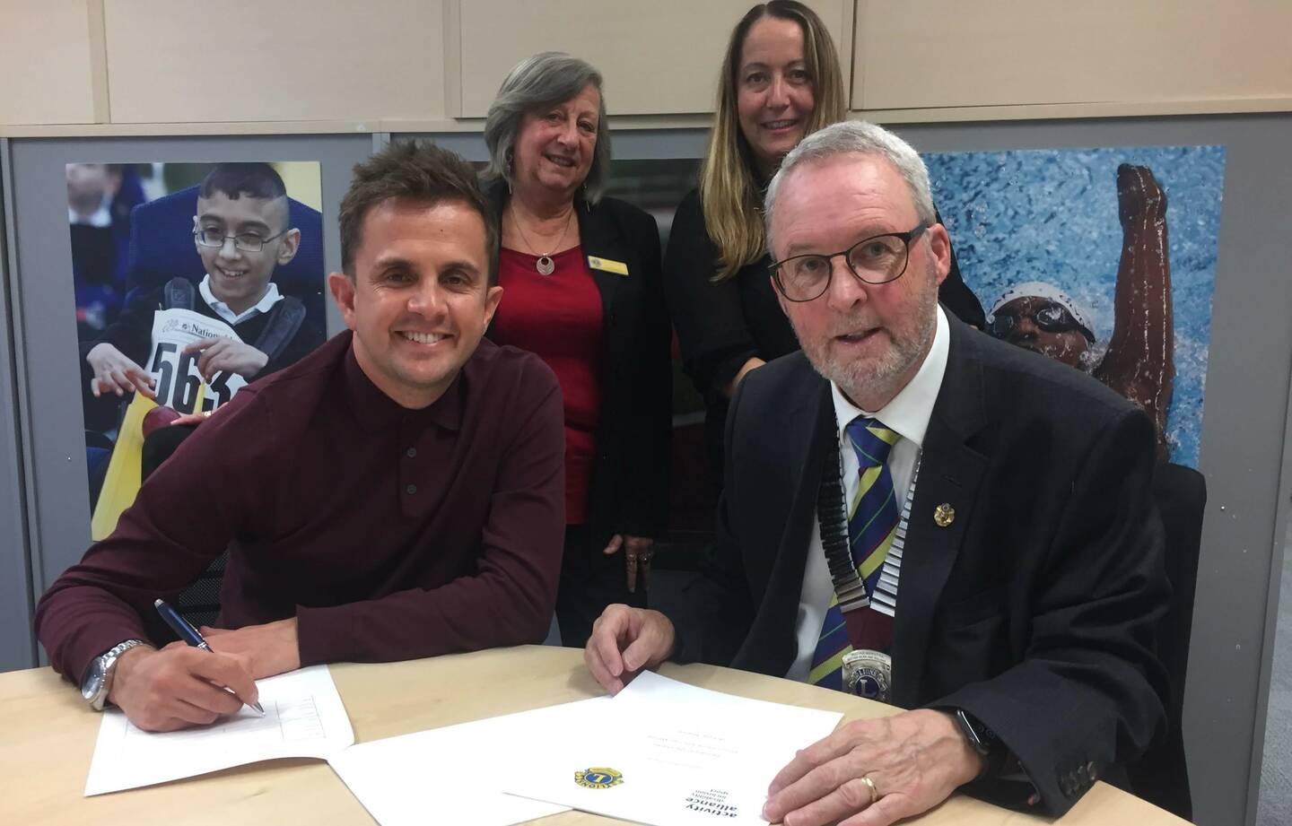 Two seated men sign pieces of paper, with two women stood behind them.