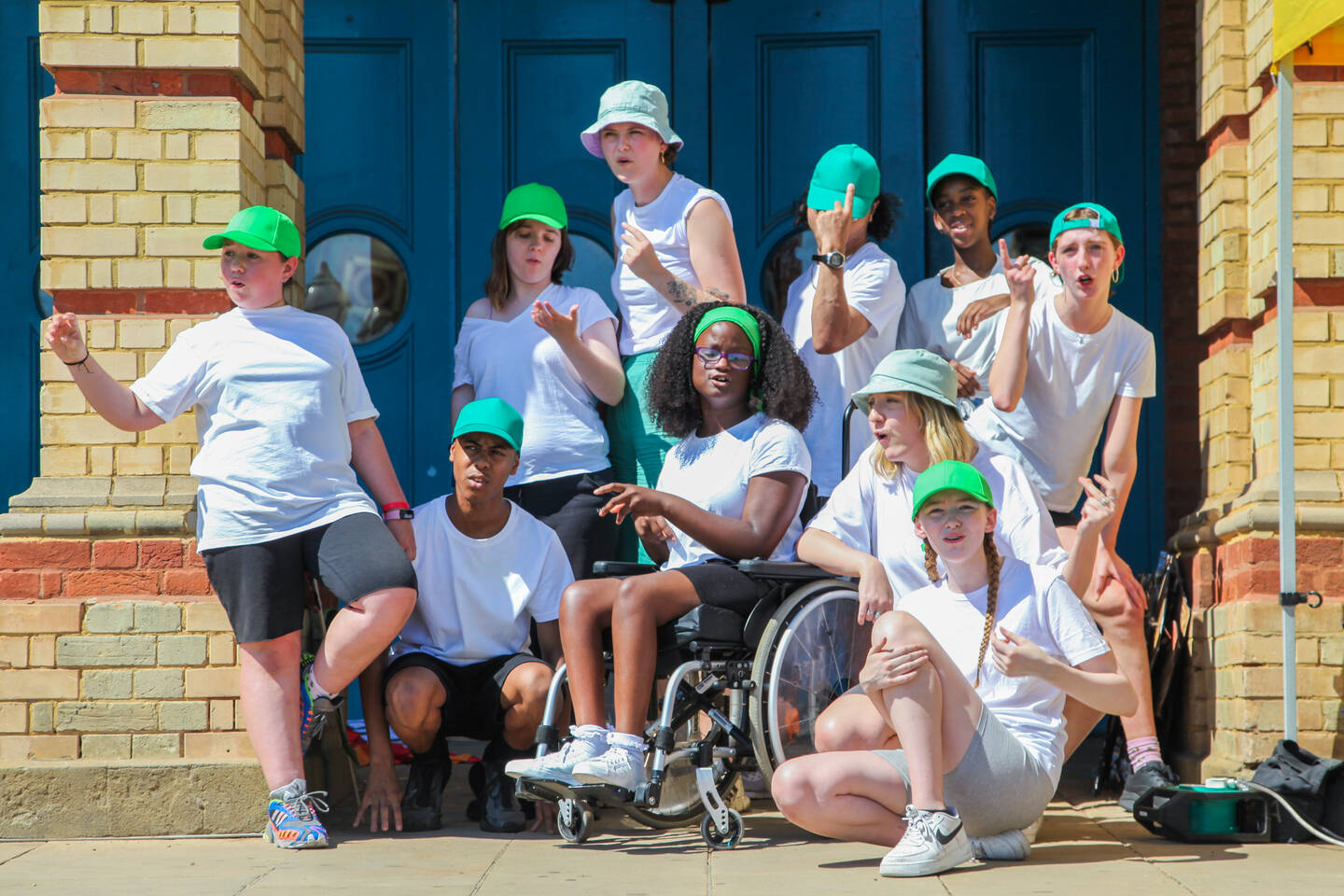 A group of children in white t-shirts and green caps stand in front of a blue door looking on.