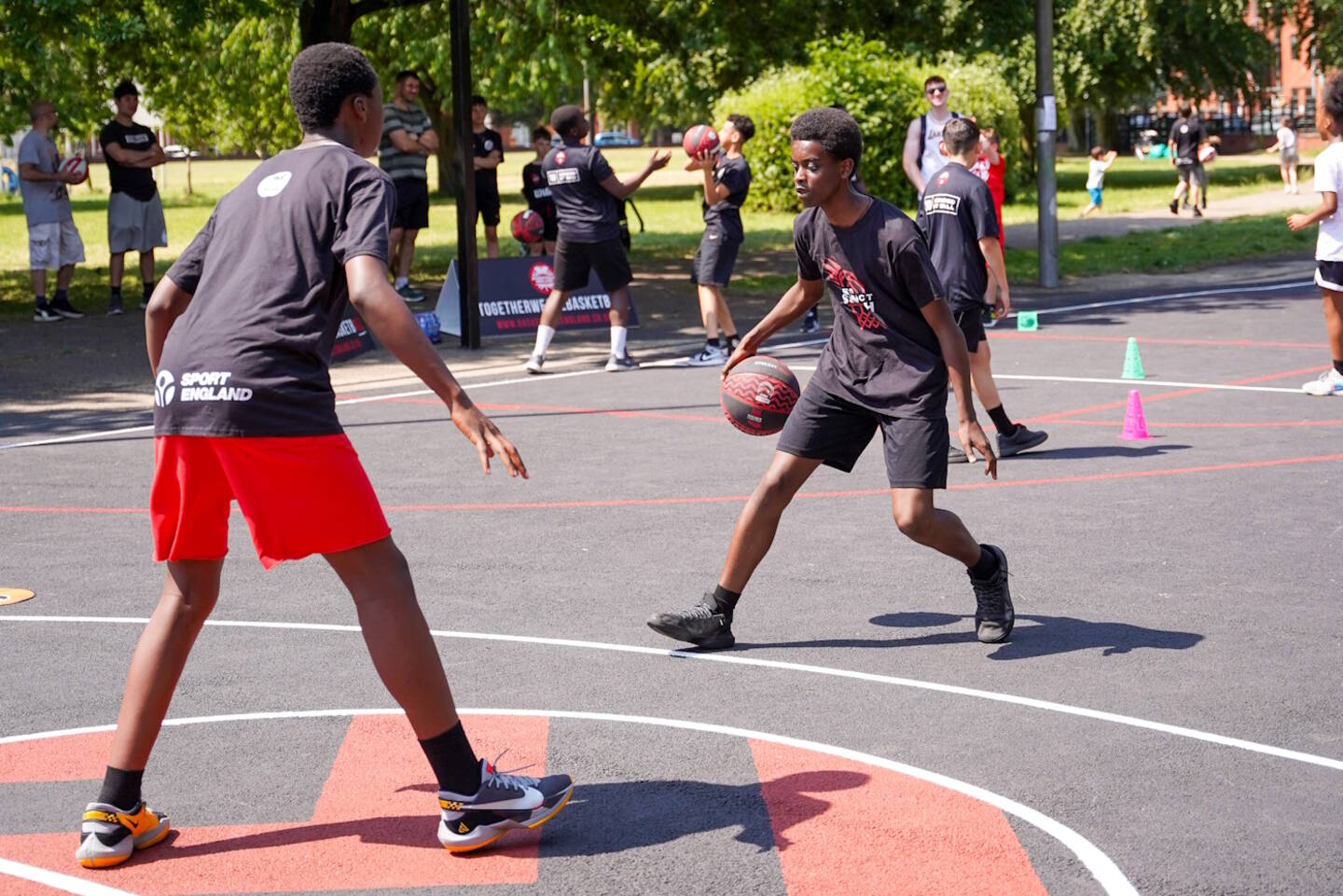 One boy dribbles a basketball towards another on an outdoor court.