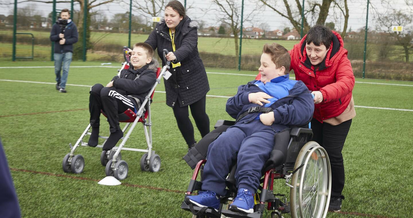Two people push two young wheelchair users across a sports field.
