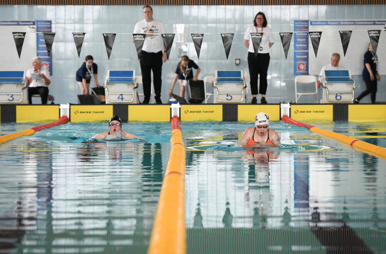 Two swimmers swim down a lane facing the camera. Officials watch them in the background.