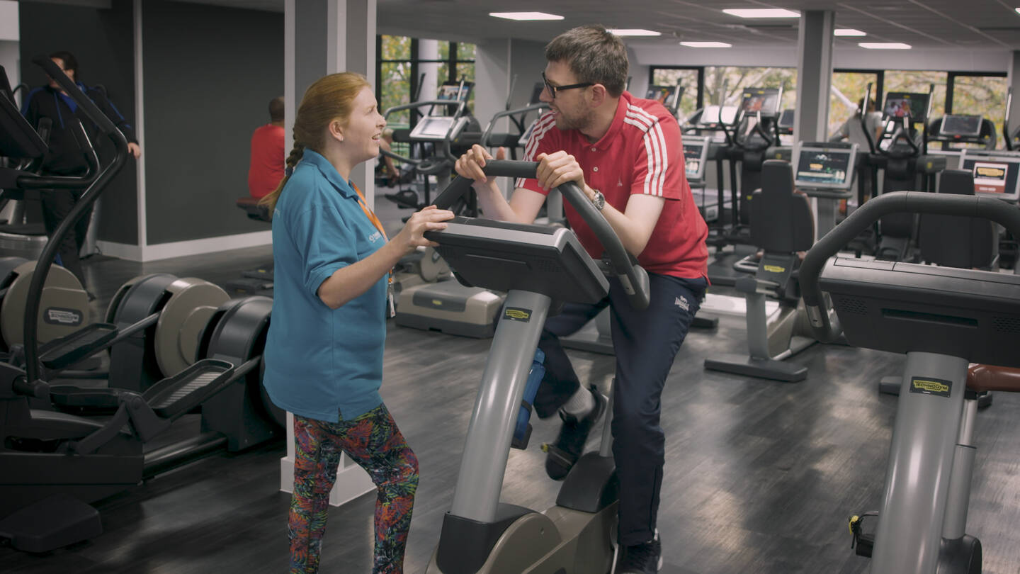 A female coach supports a man on a gym bike.