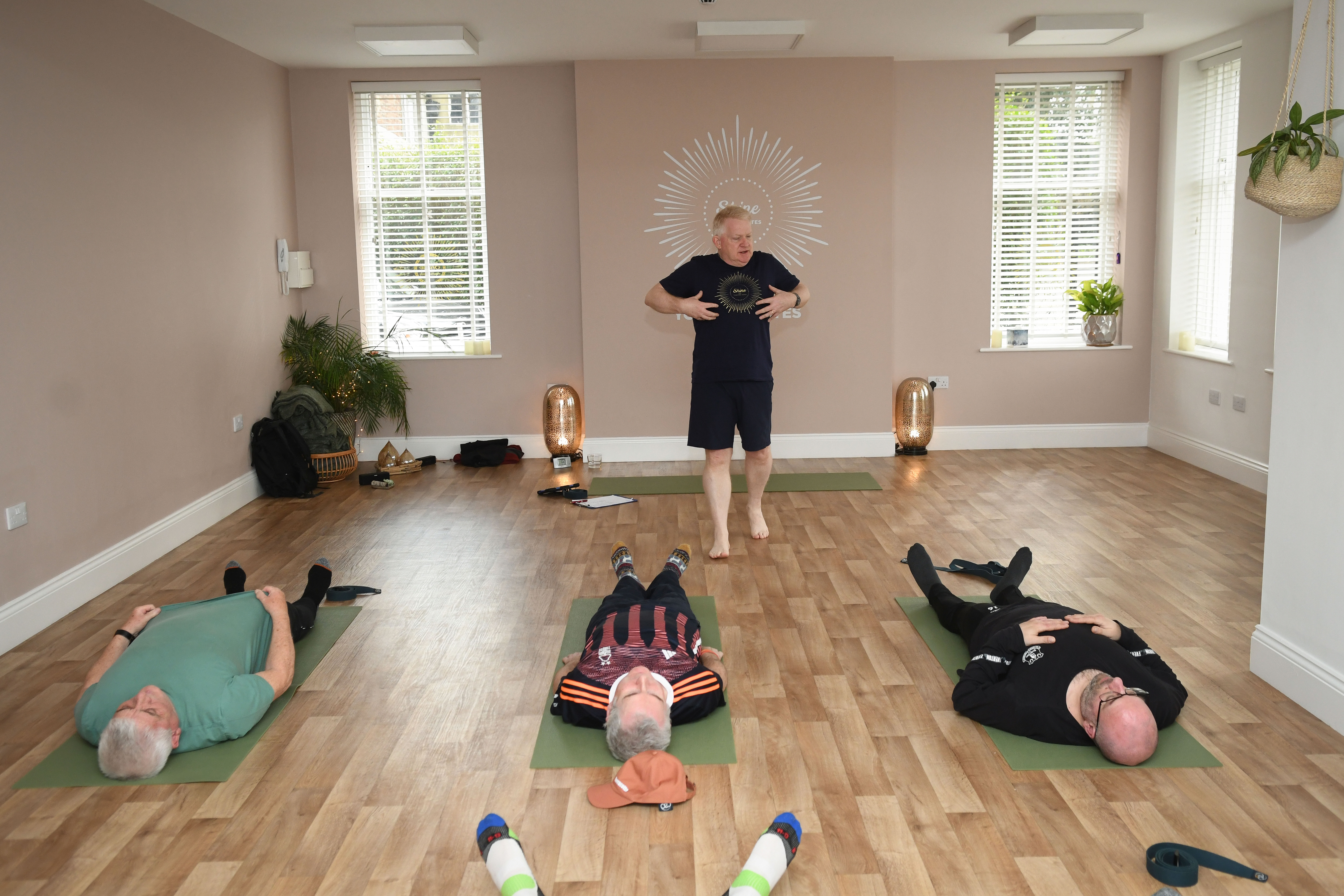 A group of men do yoga in a small sports venue. One man is stood at the front leading the session while three men lay on yoga mats.