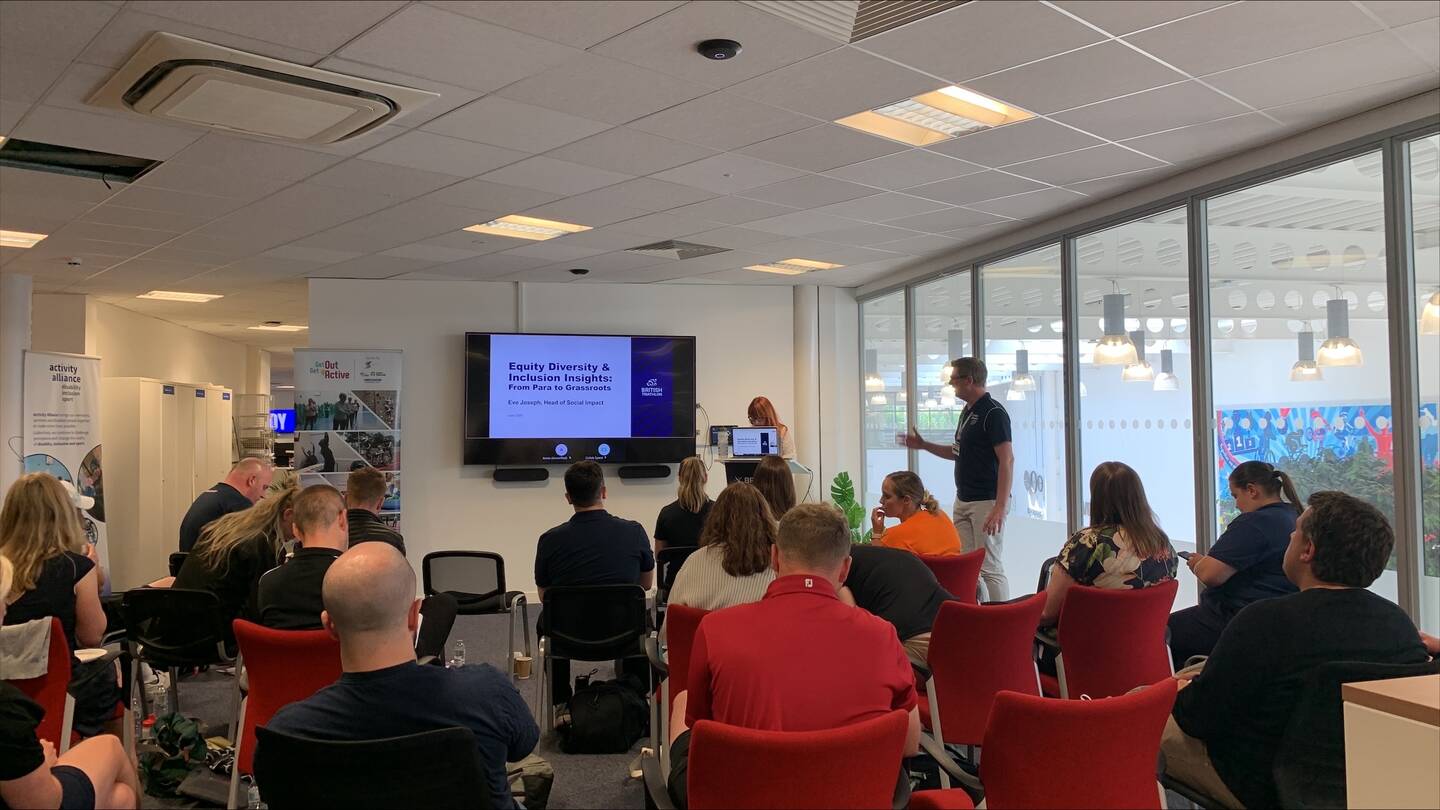 A room of people sit looking at a TV screen with two people stood at the front presenting.