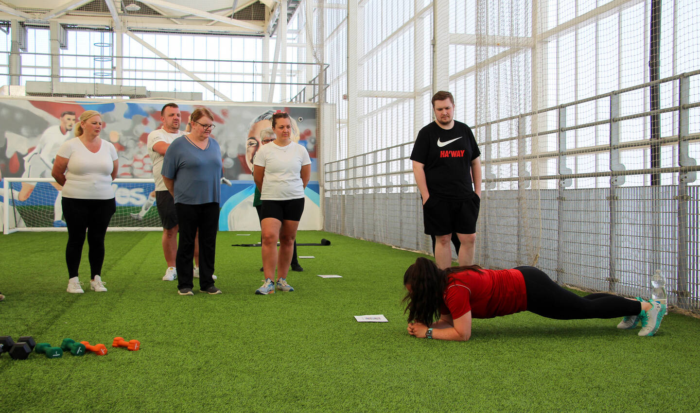 Five people stand and watch a woman do a plank at an indoor football pitch.