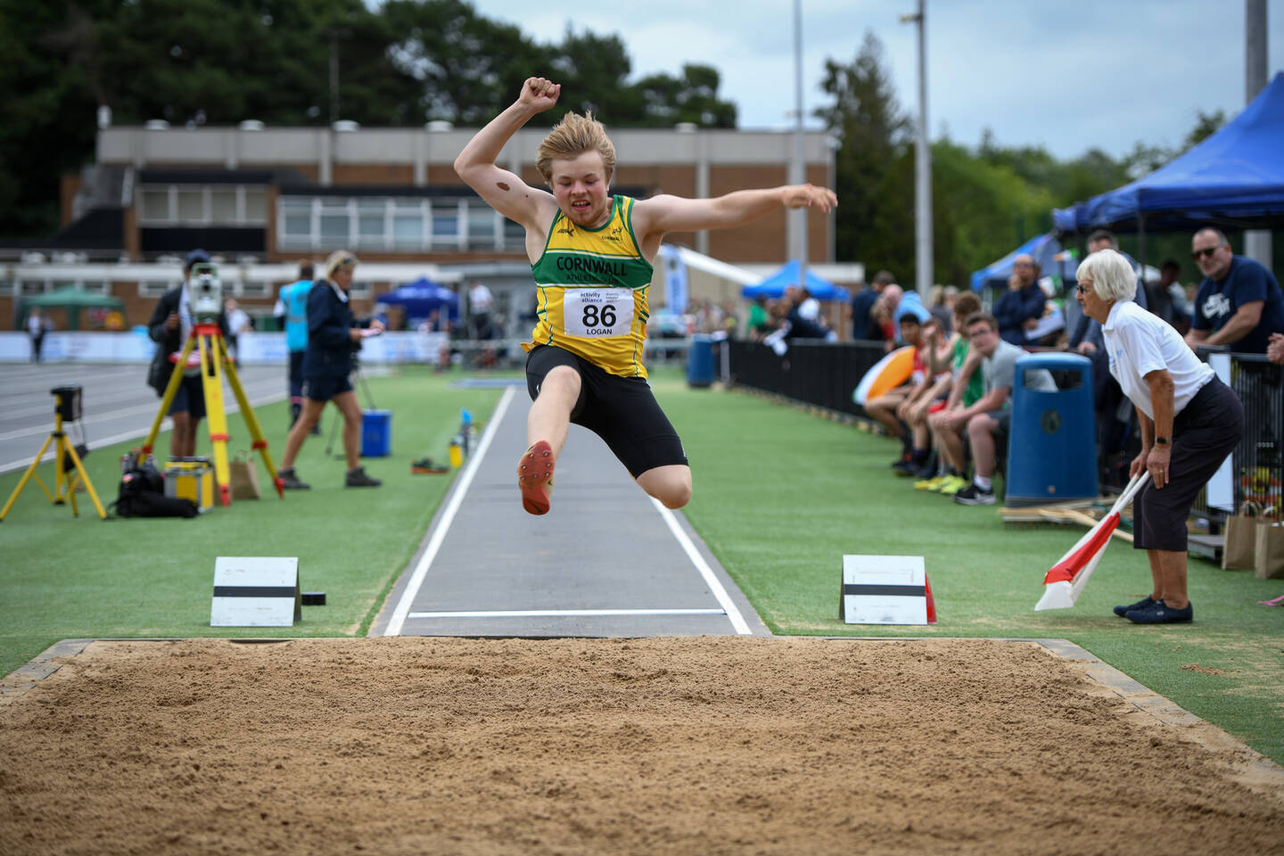 A young disabled man is photographed mid-long-jump. He is in the air about to land on the sand.