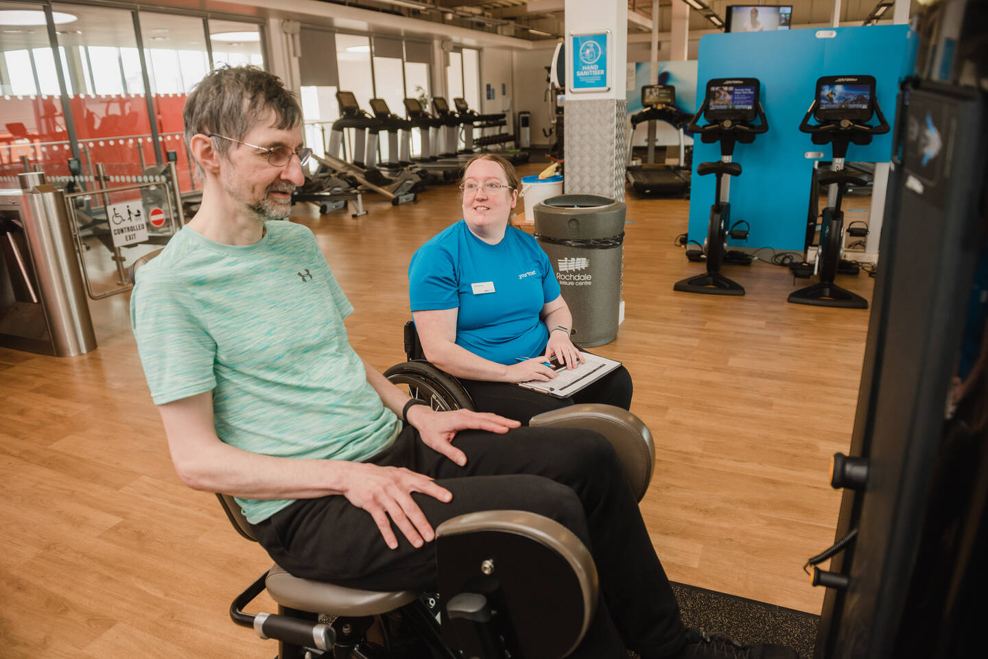 Older man in a gym using a leg weights machine. There is an instructor sat behind him.