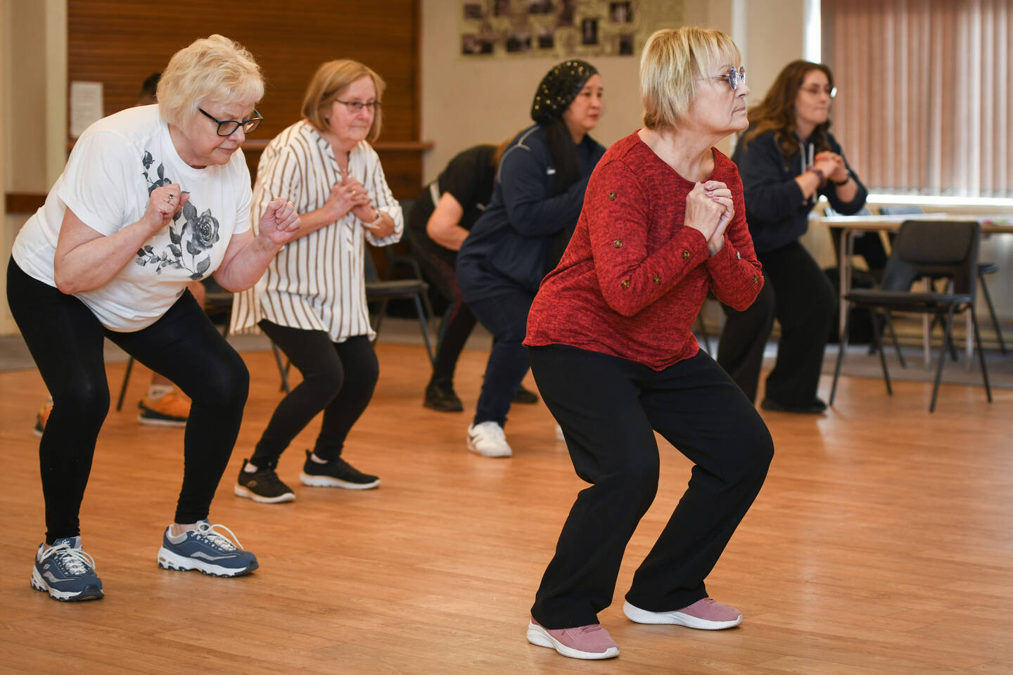 A group of older women take part in a fitness class. They are all bending their knees with hands clasped.