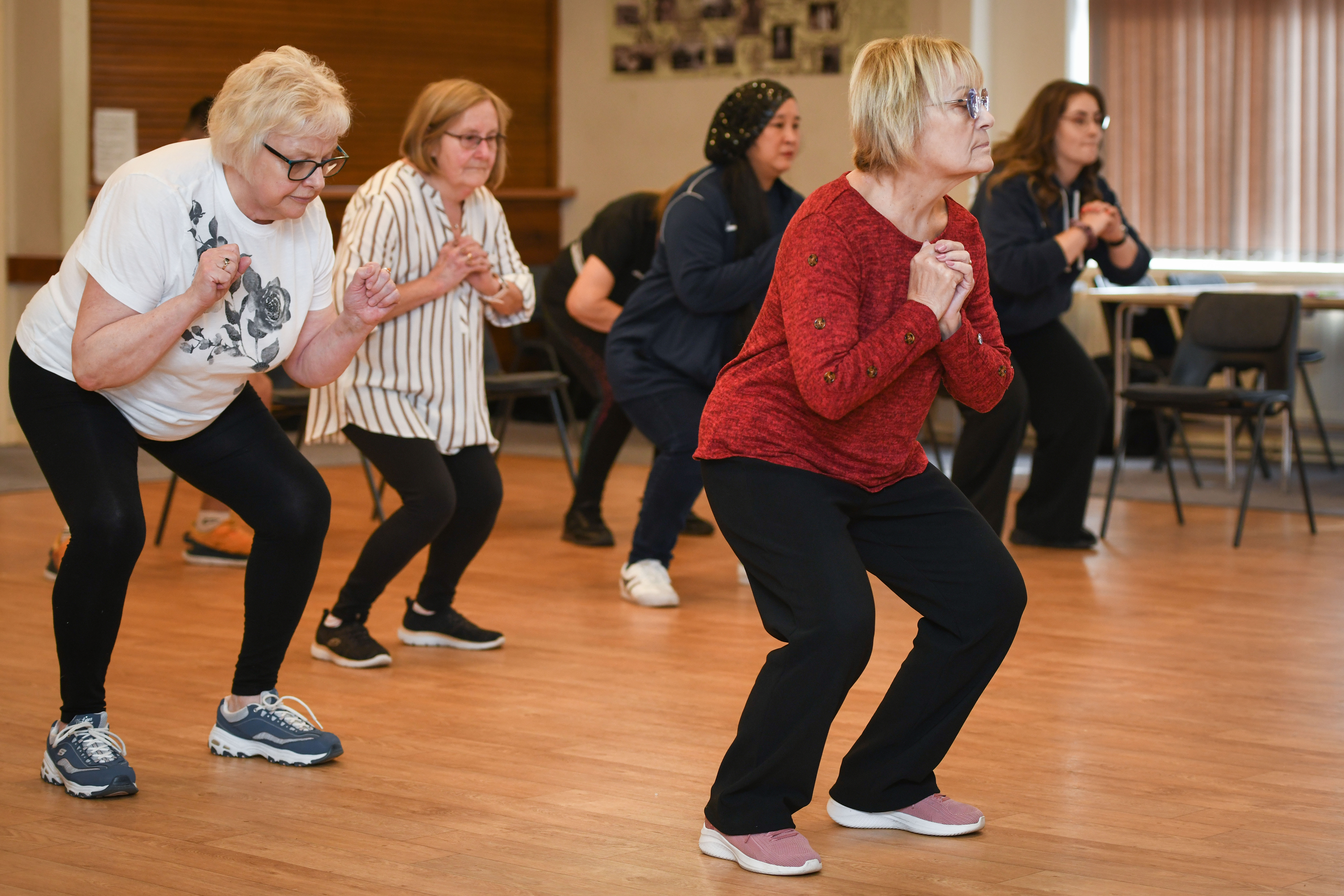 A group of older women take part in a fitness class. They are all bending their knees with hands clasped.