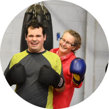 A young disabled man wears boxing gloves and stands next to a punching bag. There is a woman stood behind him smiling.
