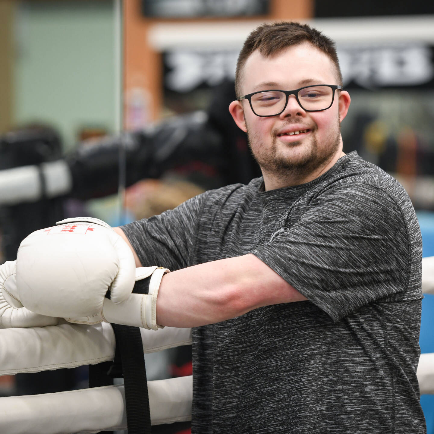 Young man with Downs Syndrome in a boxing gym. He is stood in front of the ring, smiling at the camera.