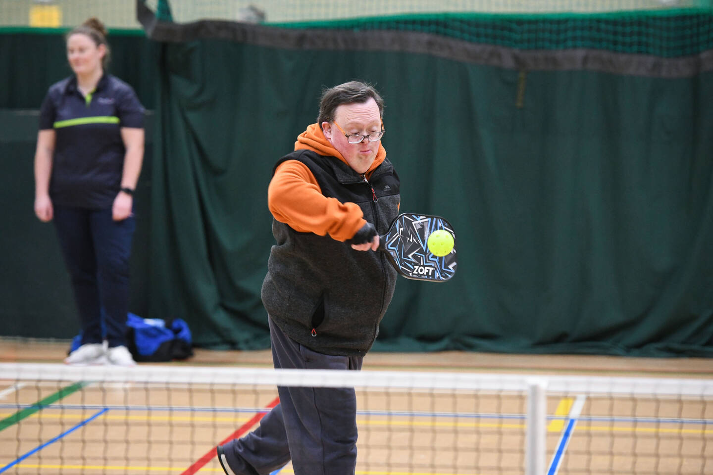A man hits a tennis ball with a small racquet over a net in an indoor sports hall.