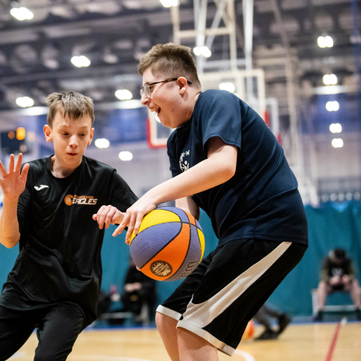 Two young disabled boys play basketball in a sports hall. One is holding the basketball while the other tried to take it off him.