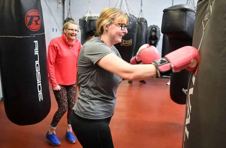 Woman boxing in a gym. She is punching a punching bag. There is a woman stood behind her looking on and smiling.