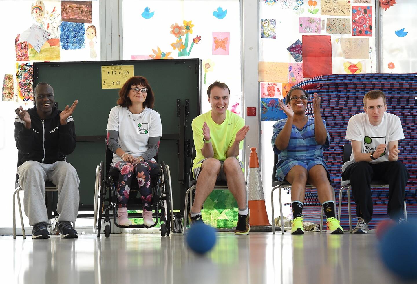 A group of disabled people sit in a row in a sports hall. They are playing a ball game.