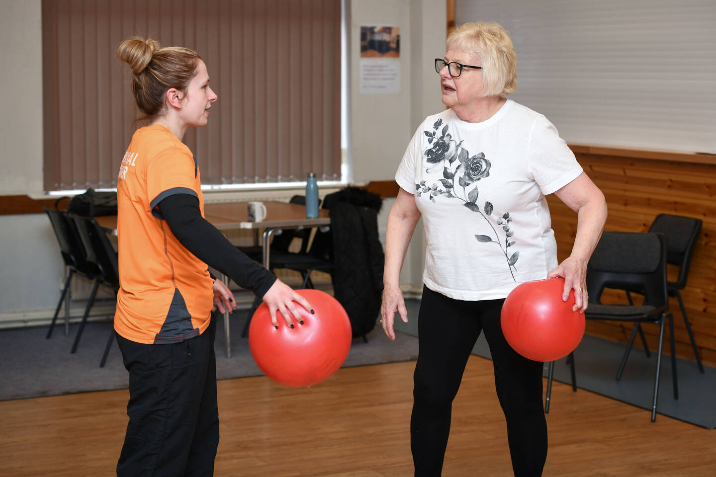 An older woman and a young woman both hold red balls and chat in a sports hall.
