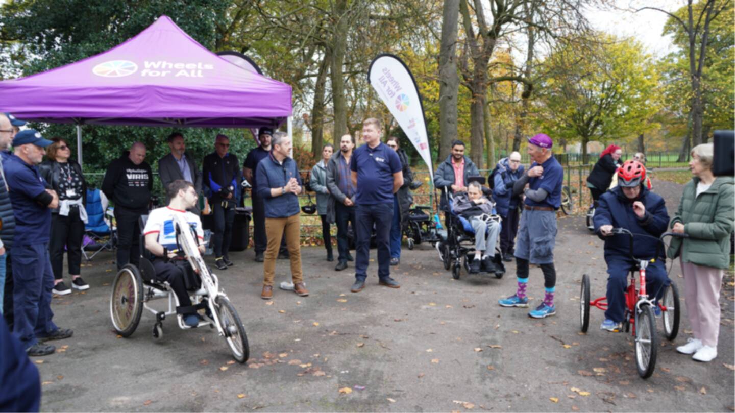 Chris Boardman stands outside with 20 people listening to him speak. They are stood around him, with a gazebo and bikes nearby.