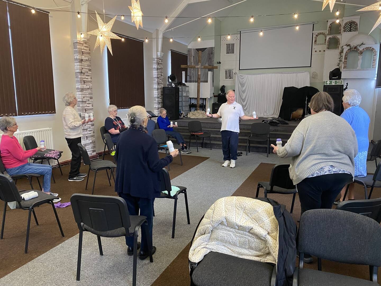 A group of elderly people do standing exercises in a church space.