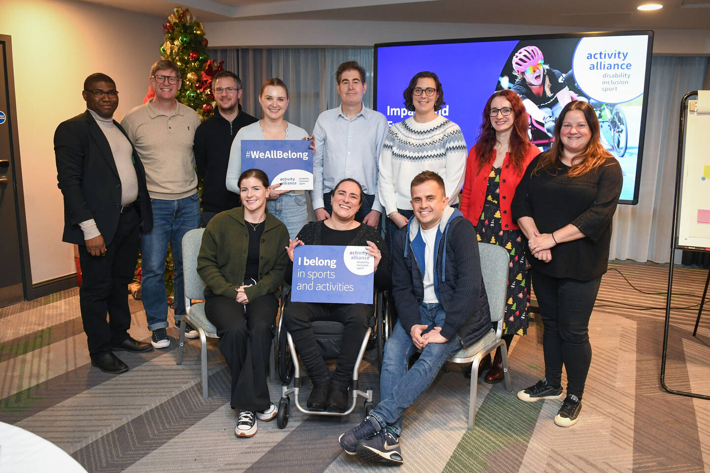 A group of Activity Alliance staff and board members stand in a room. Some are holding signs that say we all belong.