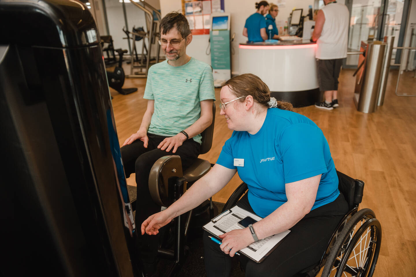 A women in a blue top using a wheelchair shows a man in a green top and glasses what to do on a gym machine.