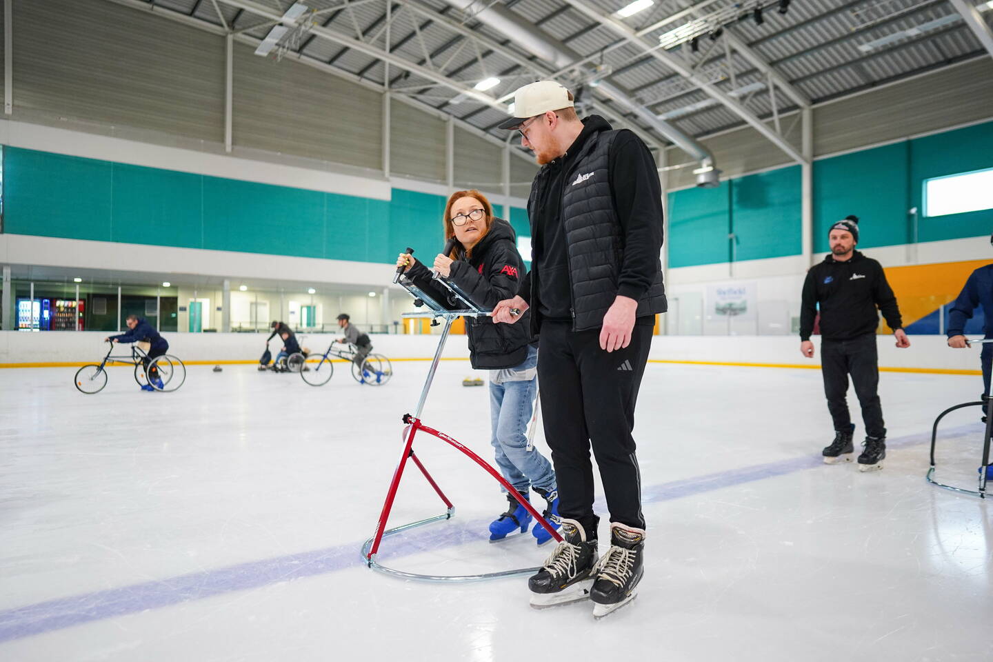 A man skates on an ice rink with a woman using a standing aid.