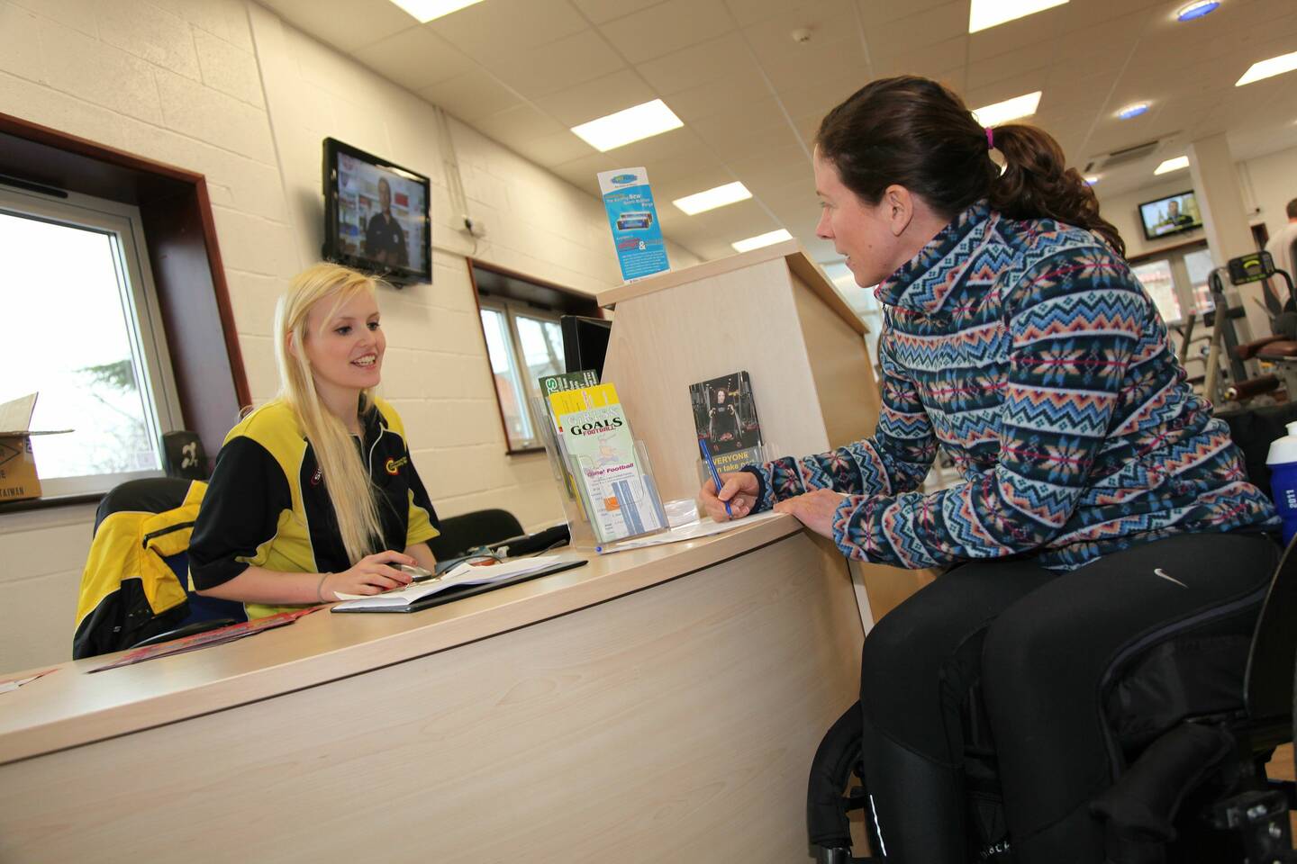 A seated woman talks to a receptionist in a gym.