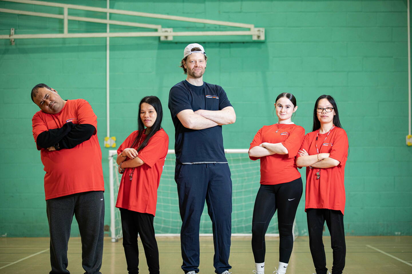 Five people pose for a photo, standing in a line smiling at the camera in a sports hall.