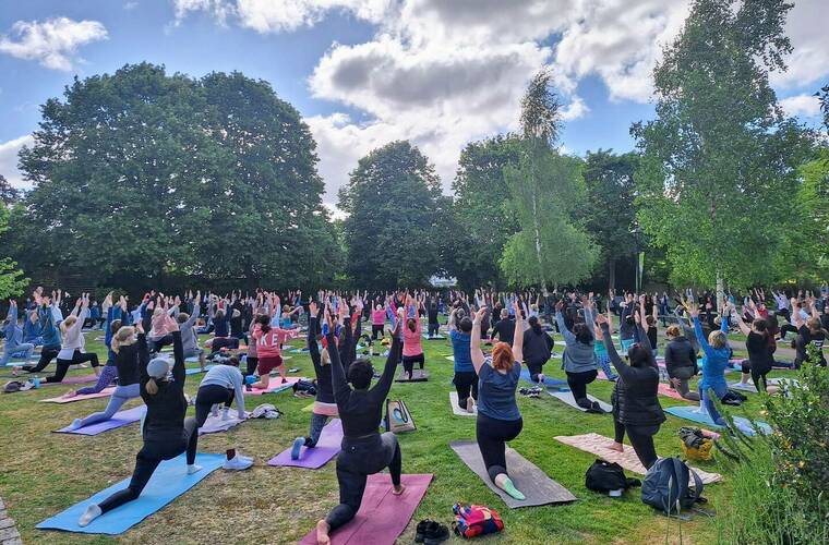 A group of people stand in a park on Yoga mats with their arms outstretched. Credit Park Yoga