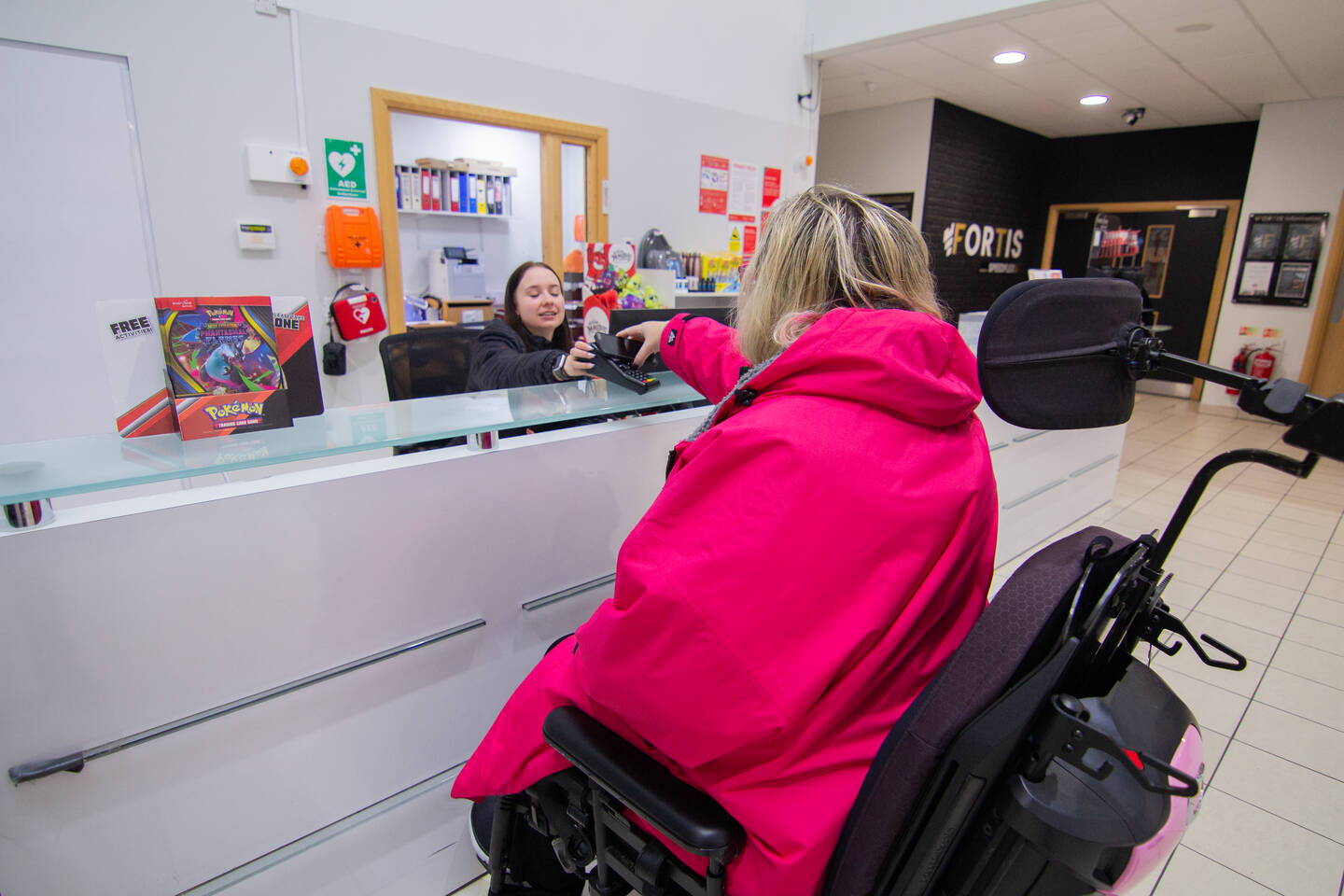 A woman behind a customer service desk takes a card payment from a woman using a wheelchair.