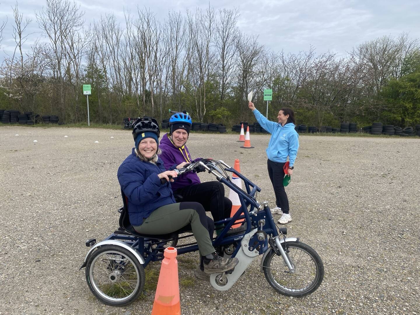 Two people sit on an adapted bike smiling as they ride in a tarmaced area.