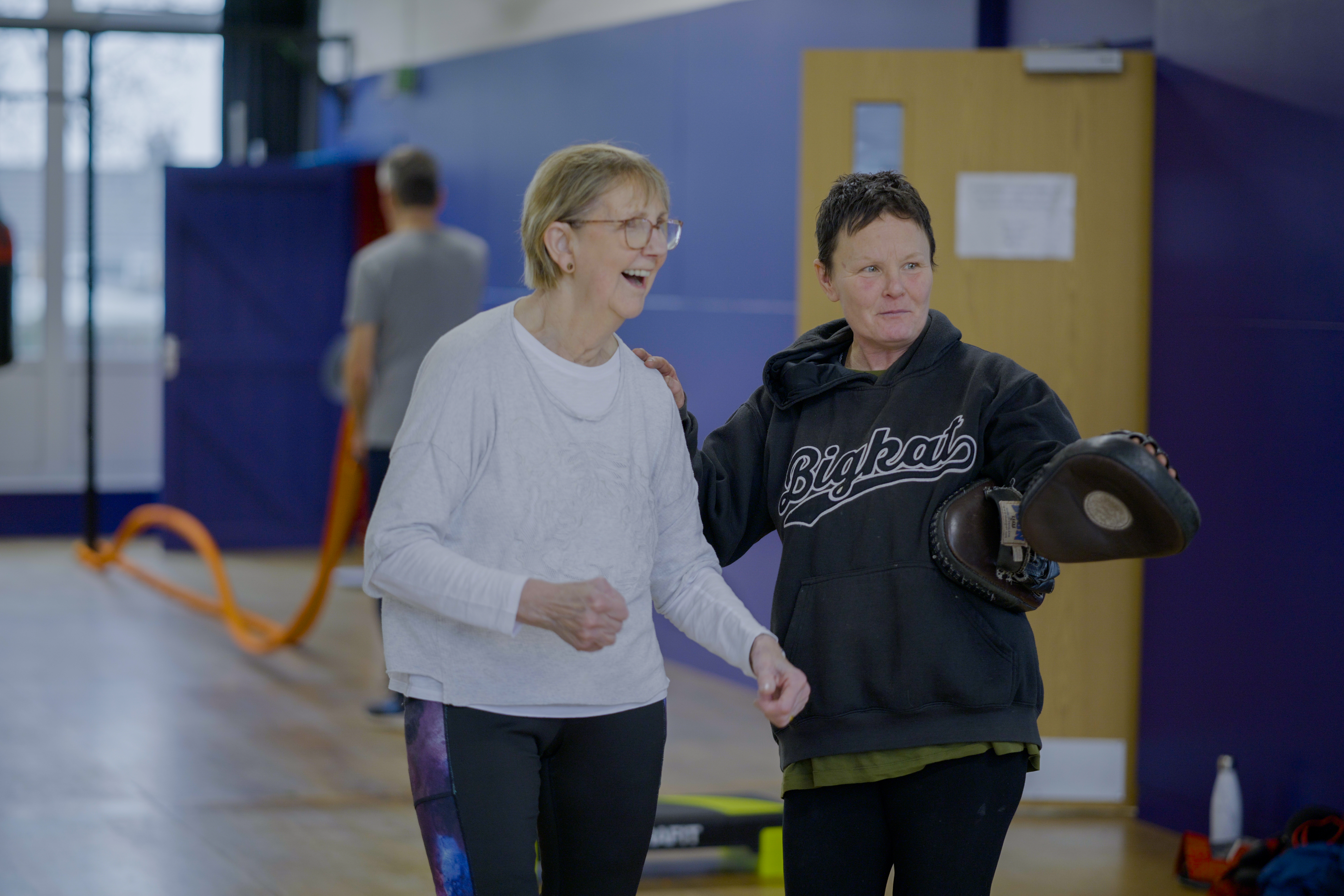 A woman smiles whilst holding a boxing pad. She is pointing at something alongside another woman.