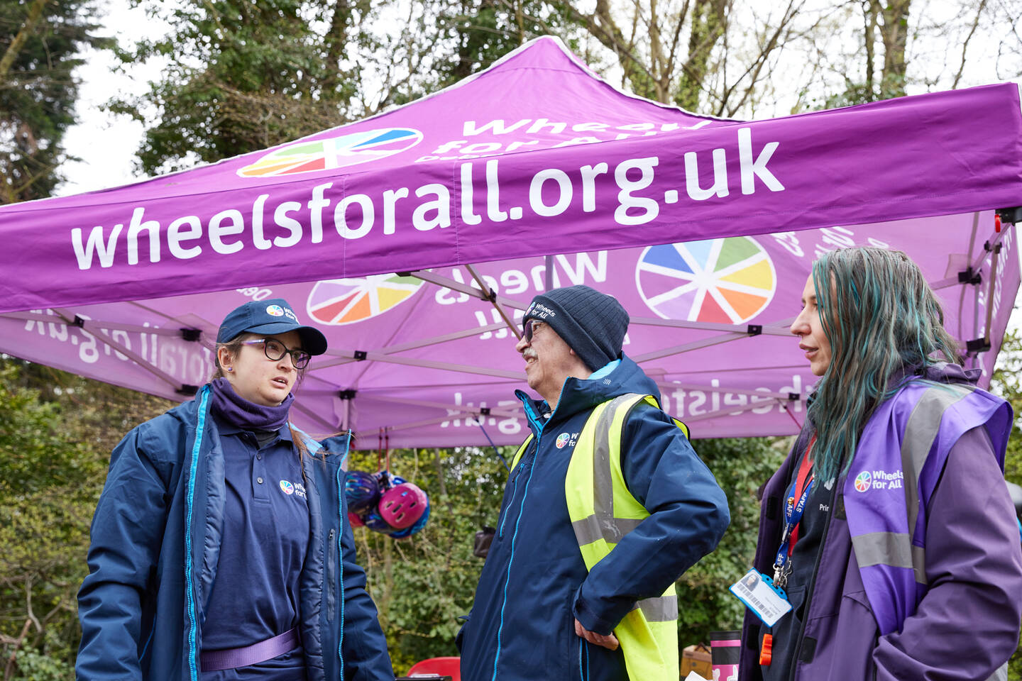 Three people stand in front of a tent with information about Wheels for All on it.