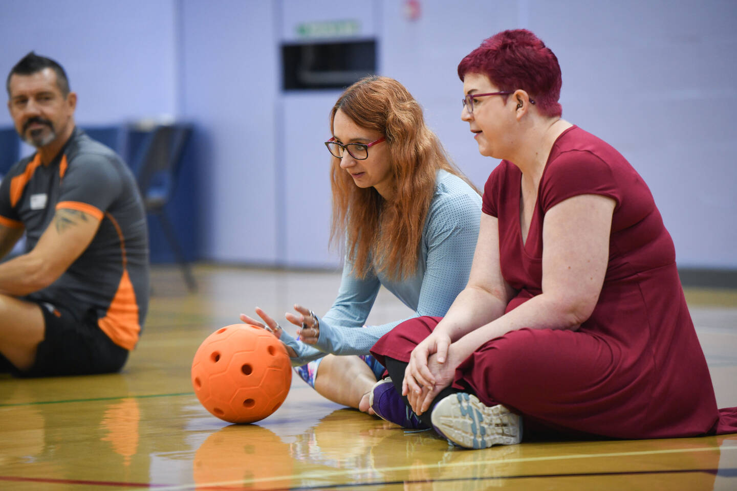Two women sit in a sports hall and roll a ball.